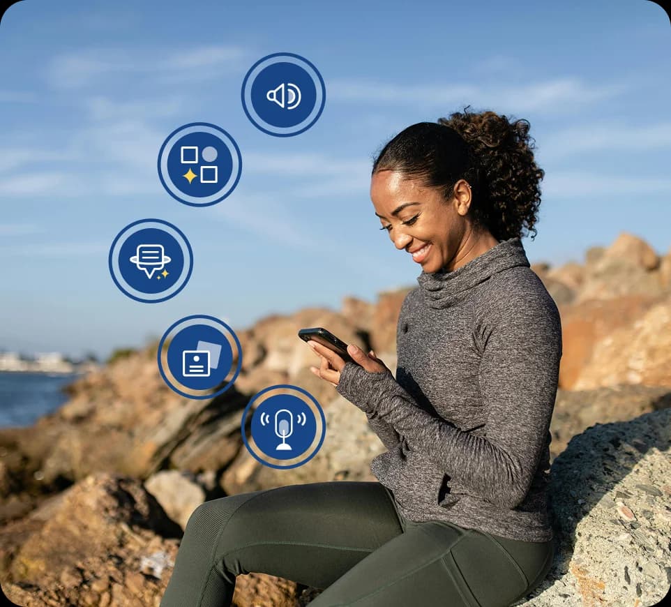 Une femme assise sur des rochers au bord de l'eau sourit en regardant son téléphone, avec des icônes représentant les fonctionnalités de Rosetta Stone Sapphire affichées autour d'elle.